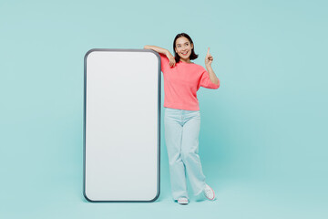 Full body young woman of Asian ethnicity in pink sweater stand near big mobile cell phone with blank screen workspace area point finger up with new idea isolated on pastel plain light blue background.