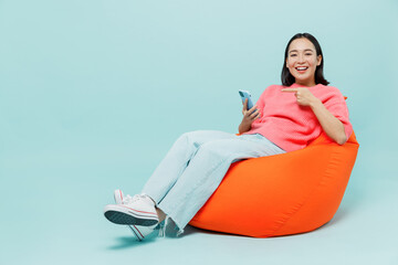 Full body young smiling fun woman of Asian ethnicity 20s wear pink sweater sit in bag chair use point finger on mobile cell phone isolated on pastel plain light blue color background studio portrait.