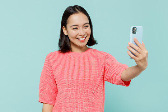 Young smiling happy woman of Asian ethnicity 20s wearing pink sweater doing selfie shot on mobile cell phone post photo on social network isolated on pastel plain light blue color background studio