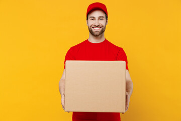 Professional happy delivery guy employee man 20s in red cap T-shirt uniform workwear work as dealer courier hold cardboard box isolated on plain yellow background studio portrait. Service concept - obrazy, fototapety, plakaty