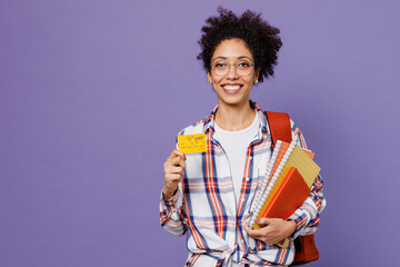 Young happy girl woman of African American ethnicity teen student in shirt backpack hold in hand books credit bank card isolated on plain purple background Education in high school university concept