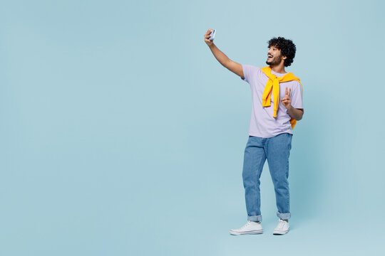 Full size young bearded Indian man 20s years old wears white t-shirt doing selfie shot on mobile cell phone post photo on social network isolated on plain pastel light blue background studio portrait.