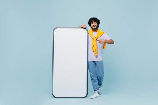 Full size young bearded Indian man 20s wears white t-shirt stand near pointing on big mobile cell phone with blank screen workspace area isolated on plain pastel light blue background studio portrait.