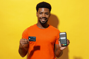 Young man of African American ethnicity 20s wear orange t-shirt hold wireless modern bank payment terminal to process acquire credit card payments isolated on plain yellow background studio portrait - obrazy, fototapety, plakaty