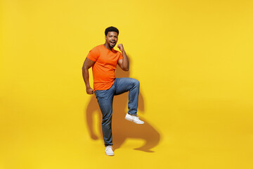 Fototapeta na wymiar Full body happy young man of African American ethnicity 20s wear orange t-shirt doing winner gesture celebrate clenching fists say yes raise up leg isolated on plain yellow background studio portrait
