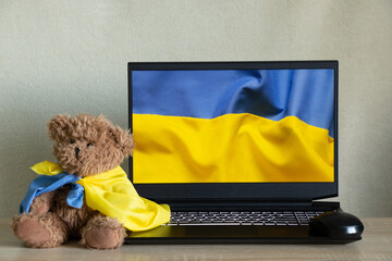 The national flag of Ukraine as a background on a laptop and a teddy bear sits at home next to the...