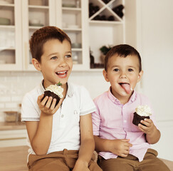 Funny two brother male kids are sitting together at the kitchen table and eating cupcakes.