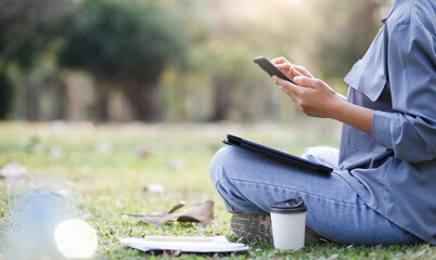Young asian woman sitting and relaxing on grass she using mobile phone working online and put tablet computer on her leg with paperwork and coffee cup on grass