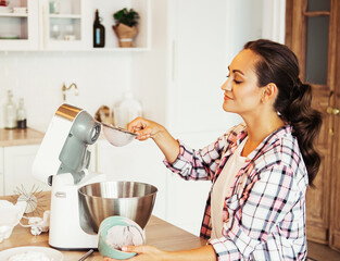 beautiful girl preparing dough in a food processor while standing in a bright kitchen