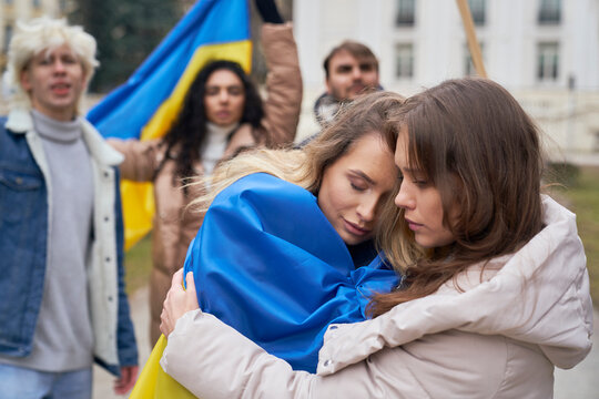 Two young caucasian women embracing in front and group of people manifesting against war in Ukraine in the background