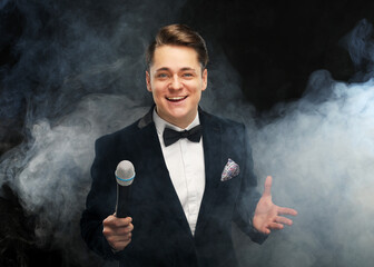 Stylish young man in a tuxedo holding a microphone, posing against a dark background with smoke
