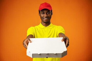 Smiling black male courier wearing uniform holding box in orange studio