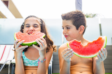 Two funny kids eating watermelon at the the edge of the pool on a summer day