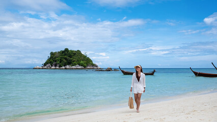 Obraz premium Portrait image of a beautiful young asian woman with hat and bag strolling on the beach with the sea and blue sky background