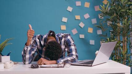 Overworked man putting head on desk and showing okay sign, giving thumbs up while he feels exhausted. Tired and sleepy employee advertising good and positive gesture with hands.