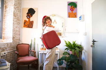 Young, beautiful, bohemian Latina, wearing a red beret on her head and a red vintage suitcase, looking happily at the camera for the photo. Concept of an enterprising woman.