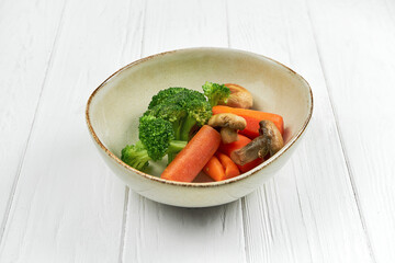 Boiled vegetables (broccoli, carrots, mushrooms) in a plate on a white wooden background.