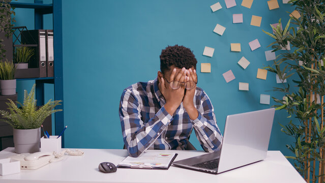 Desperate employee working on laptop computer at desk, feeling tired and stressed about deadline. Overworked man feeling frustrated and disappointed about job mistake. Sad person