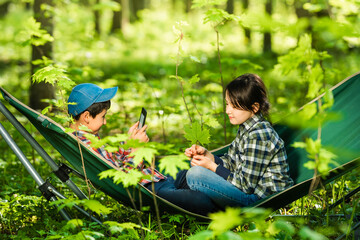 Naklejka na ściany i meble Two children boy and girl reading an e-book siting in a folding hammock in the spring or summer forest. Lots of greenery and flowers around. Camping outdoors. Harmony with nature.