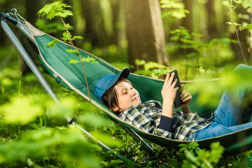 A school-age girl spends time in the nature on a spring folding hammock. The child reads an e-book, teaches lessons, learns, distance learning, harmony with nature, rest and relaxation.