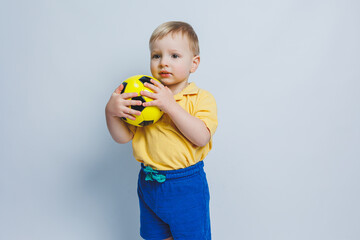 A little boy in a yellow t-shirt with a soccer ball in his hand smiles isolated on a white background. Sports child holding a ball. Children's sports game. Little athlete.