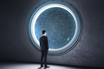 Back view of young businessman looking out of round illuminator with starry sky cosmos view in...