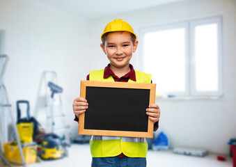 building, construction and profession concept - little boy in protective helmet and safety vest holding chalkboard over room with working equipment background