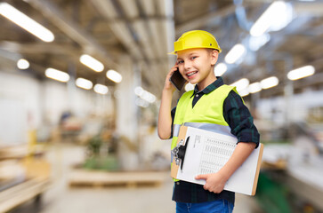 industry, manufacture and profession concept - happy smiling little boy in protective helmet and safety vest with clipboard calling on smartphone over workshop background