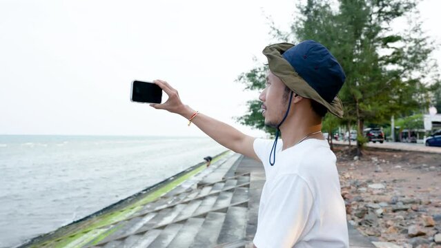 4K 25fps Slow Motion, A good-looking Asian guy, wearing a white shirt and his favorite hat, is using phone camera pictures of the sea in the evening when the sky is clear, popular tourist destination.