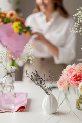 people, gardening and floral design concept - lavender flowers in vase on table over happy smiling woman or floral artist with bunch of flowers working at studio