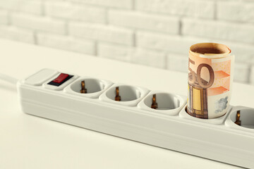 Extension board with rolled euro banknotes on white table near brick wall, closeup