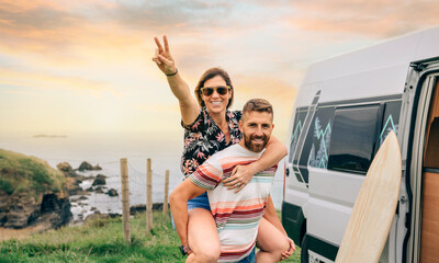 Happy young couple looking camera having fun piggybacking next to their camper van during a trip