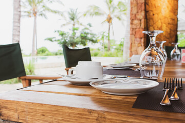Table setting in tropical resort. Table set near resort pool. Cutlery on table of outdoor restaurant. Wine glass and cup on dinner table in luxury villa. Restaurant near swimmimg pool with palm trees.