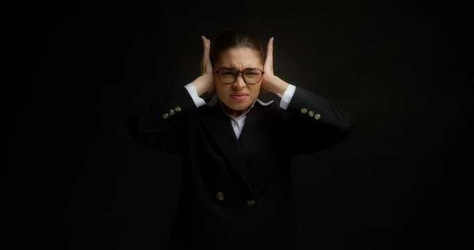 Young Asian woman in glasses and office clothes plugging ears with palms of hands from a too loud sound.A business woman is prevented from working by loud conversations on an isolated black background