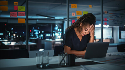 Overworked African American Businesswoman Working on Laptop Computer in Big City Office Late in the Evening. Tired Stressed Female Entrepreneur trying to Find Solution for Business Problems. 