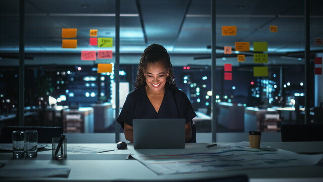 Portrait Of African American Businesswoman Working On Laptop Computer In Big City Office Late In The Evening. Female Executive Director Managing Digital E-Commerce Project, Finance Analysis