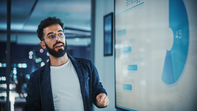 Company Creative Manager Holds Sales Meeting Presentation for Employees and Executives. Hispanic Male Uses TV Screen with Growth Analysis, Charts, Ad Revenue. Work in Business Office.