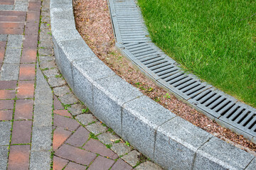 iron drainage grate on the walkway side with green turf lawn and stone pebbles at the granite curb...