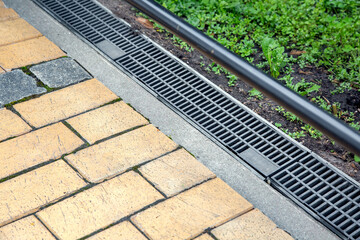iron grating of the drainage system for drainage of rainwater in the backyard at edge of sidewalk from stone brick tile with weeds in soil, landscaping close up view, nobody. - obrazy, fototapety, plakaty