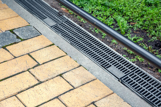 iron grating of the drainage system for drainage of rainwater in the backyard at edge of sidewalk from stone brick tile with weeds in soil, landscaping close up view, nobody.
