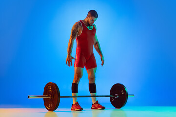 Studio shot of young man in red sportswear exercising with barbell isolated blue background in...