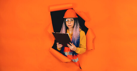 Female construction worker holding clipboard and sticking out of hole of orange background. Young woman in eyeglasses and overalls checking business project.