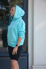 health activity. blonde girl in blue hoodie and black cycling shorts is standing in profile with hood on head near park cafe background and looking away with smile. sport lifestyle concept, free space