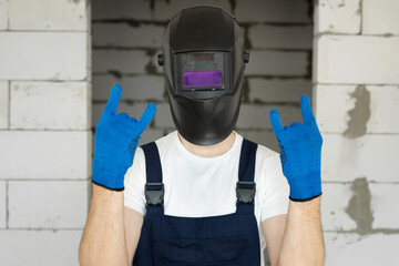 Male construction worker wearing a welding helmet and blue gloves against a brick wall.