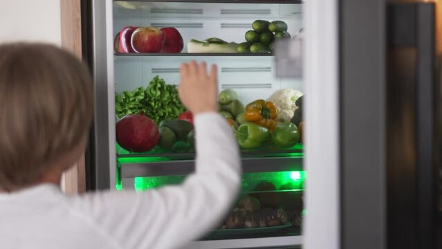 Boy opening refrigerator door while standing in kitchen. Refrigerator full of fruits and vegetables. Healthy lifestyle concept.