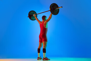 Studio shot of young man in red sportswear exercising with barbell isolated blue background in...