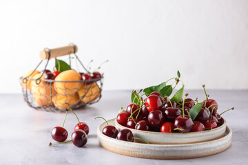 Fresh cherries in two light ceramic plates and apricots in a wicker iron basket on a gray concrete and white background.