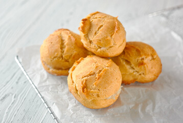 Homemade profiteroles on a metal grid and white paper on a white wooden background.
