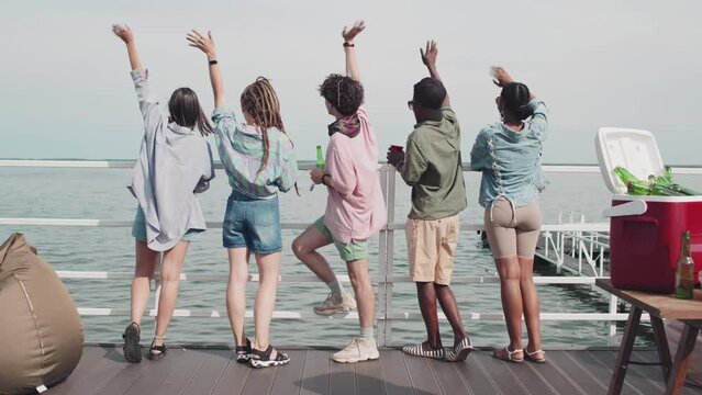 Rear of five multiethnic girls and guys standing on edge of pier on lake, drinking beer, waving hands and looking at water on sunny summer day