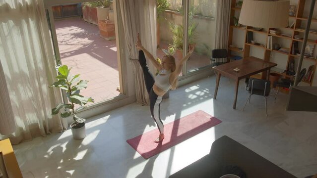 Fit, flexible blond white female performing asana sequence as daily training. In sportswear, barefoot, on the matt to stretch her body. Calm spacious interior ambient, good spring or summer weather 4k